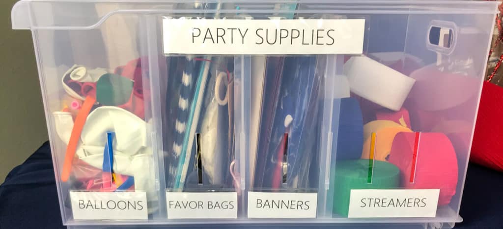 A plastic storage bin containing party supplies such as balloons, favor bags, banners, and streamers in a closet used for organizing craft supplies