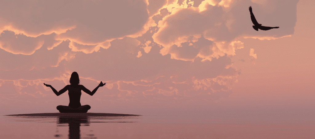 A woman meditating near water with a sunset backdrop
